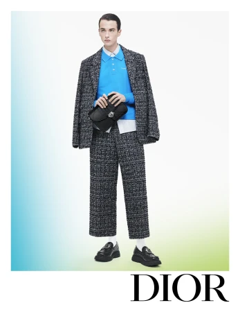 An adult male model poses in a bright studio wearing a grey speckled suit with a blue polo and black bag, with the brand name DIOR.