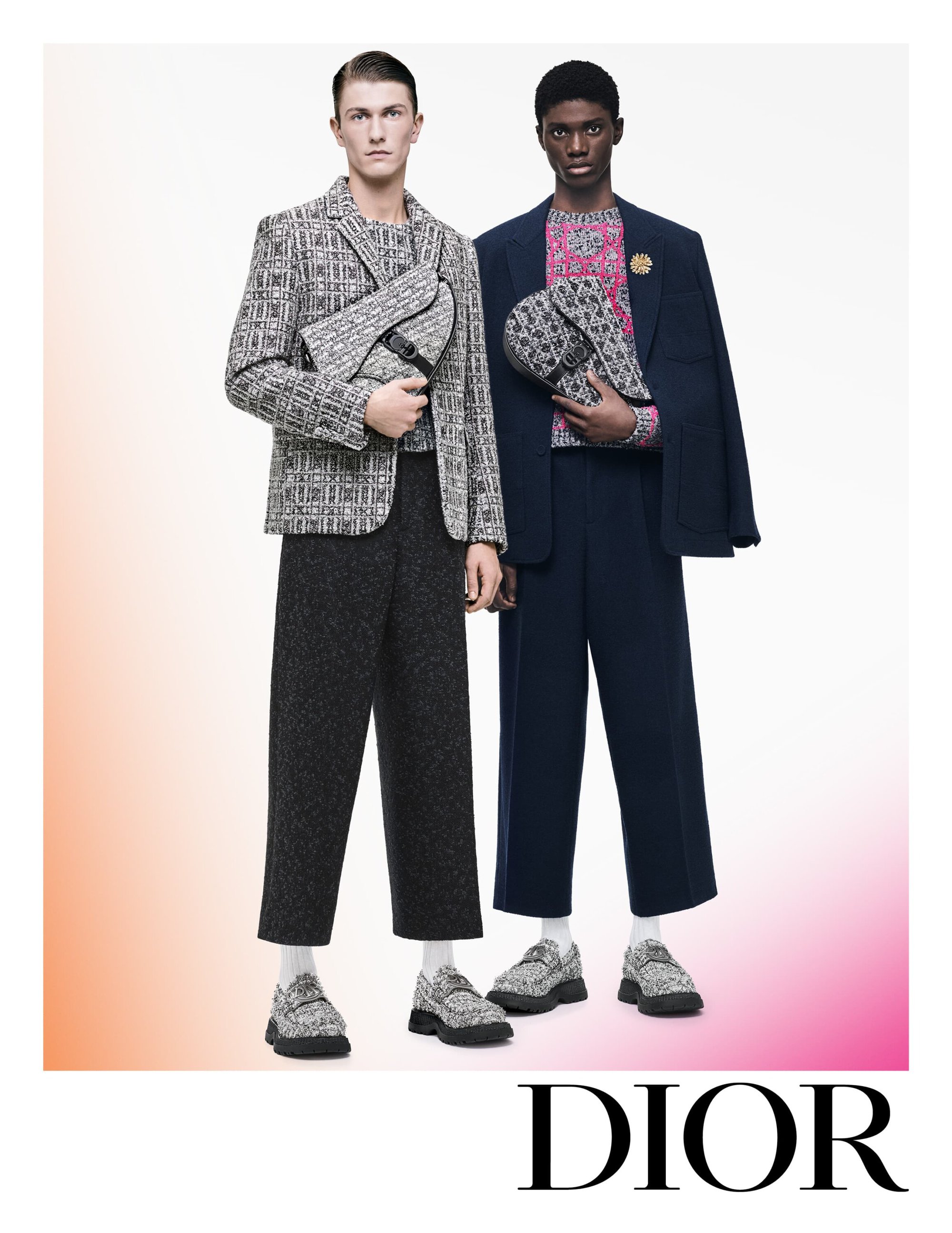 Two male fashion models pose indoors wearing patterned jackets, wide-leg trousers, and matching bags for a Dior advertisement.