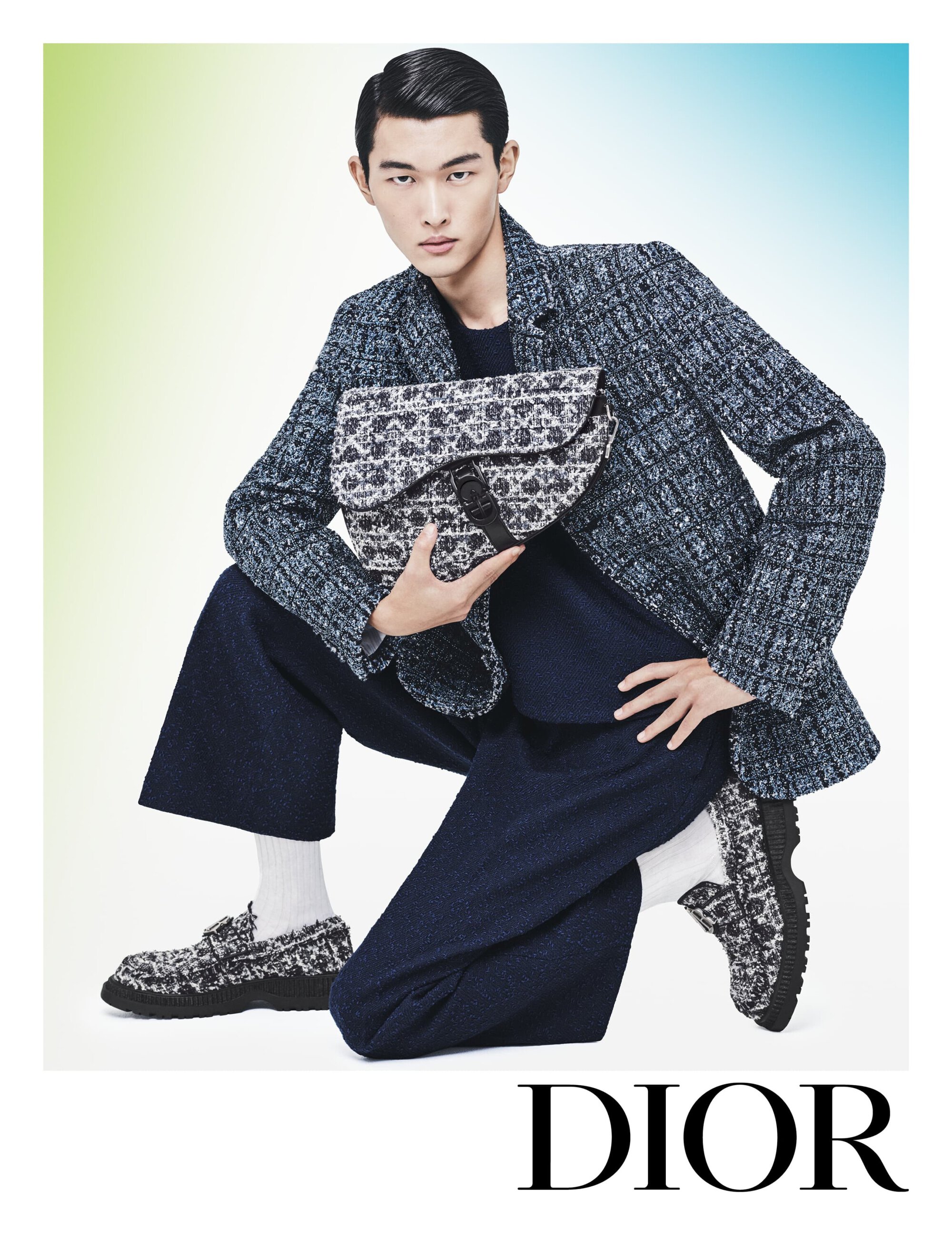 An Asian male model poses while crouching, wearing a tweed jacket, navy culottes, and loafers, holding a bag in a studio setting.