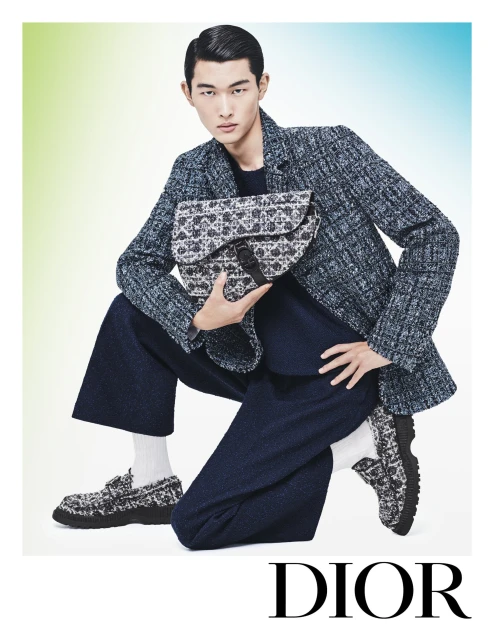 An Asian male model poses while crouching, wearing a tweed jacket, navy culottes, and loafers, holding a bag in a studio setting.
