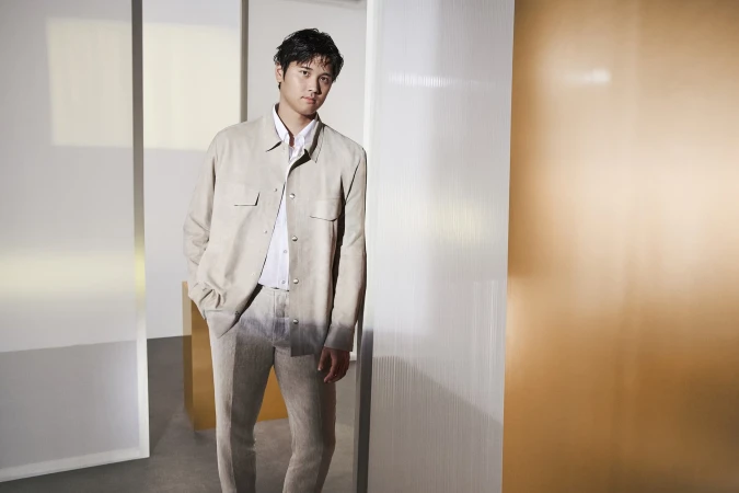 Adult male in light beige jacket and pants poses indoors against a wall, showcasing modern fashion and neutral color tones in a studio setting.