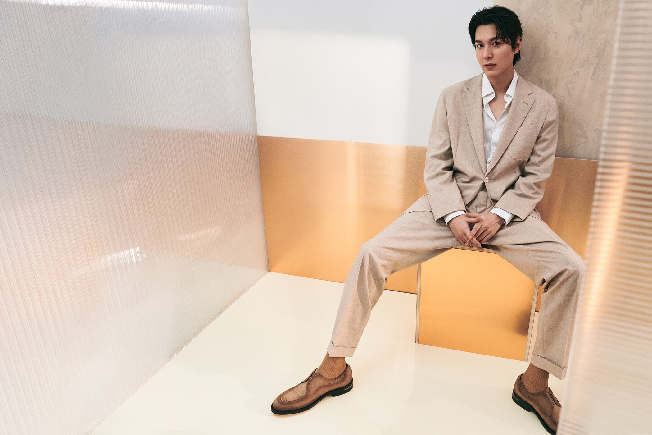 Adult male in a beige suit sits in a modern setting with brown leather shoes, posing for a portrait shot with studio lighting.