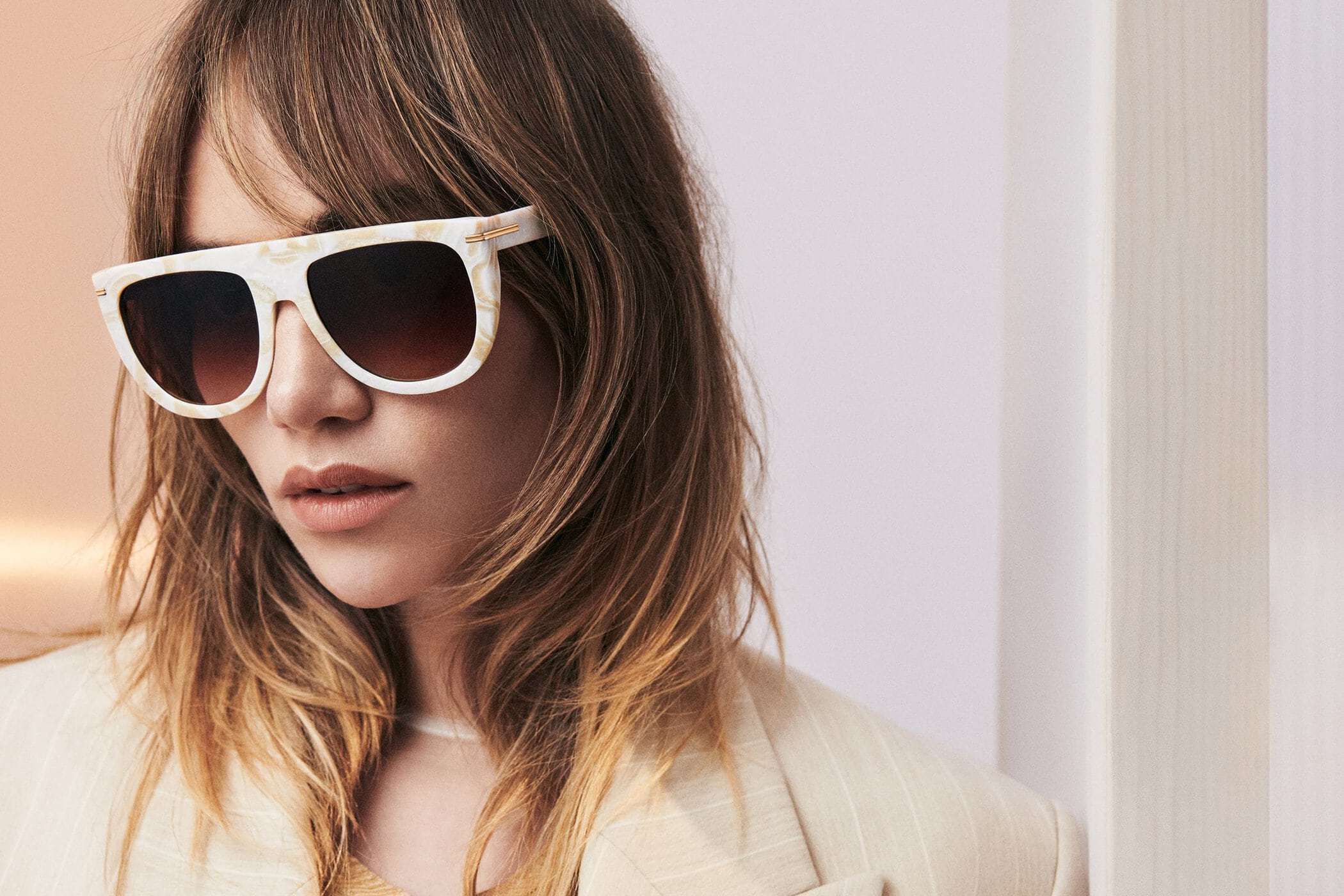 Close-up of an adult female model wearing patterned sunglasses and a blazer in a studio with a neutral background and diffused lighting.