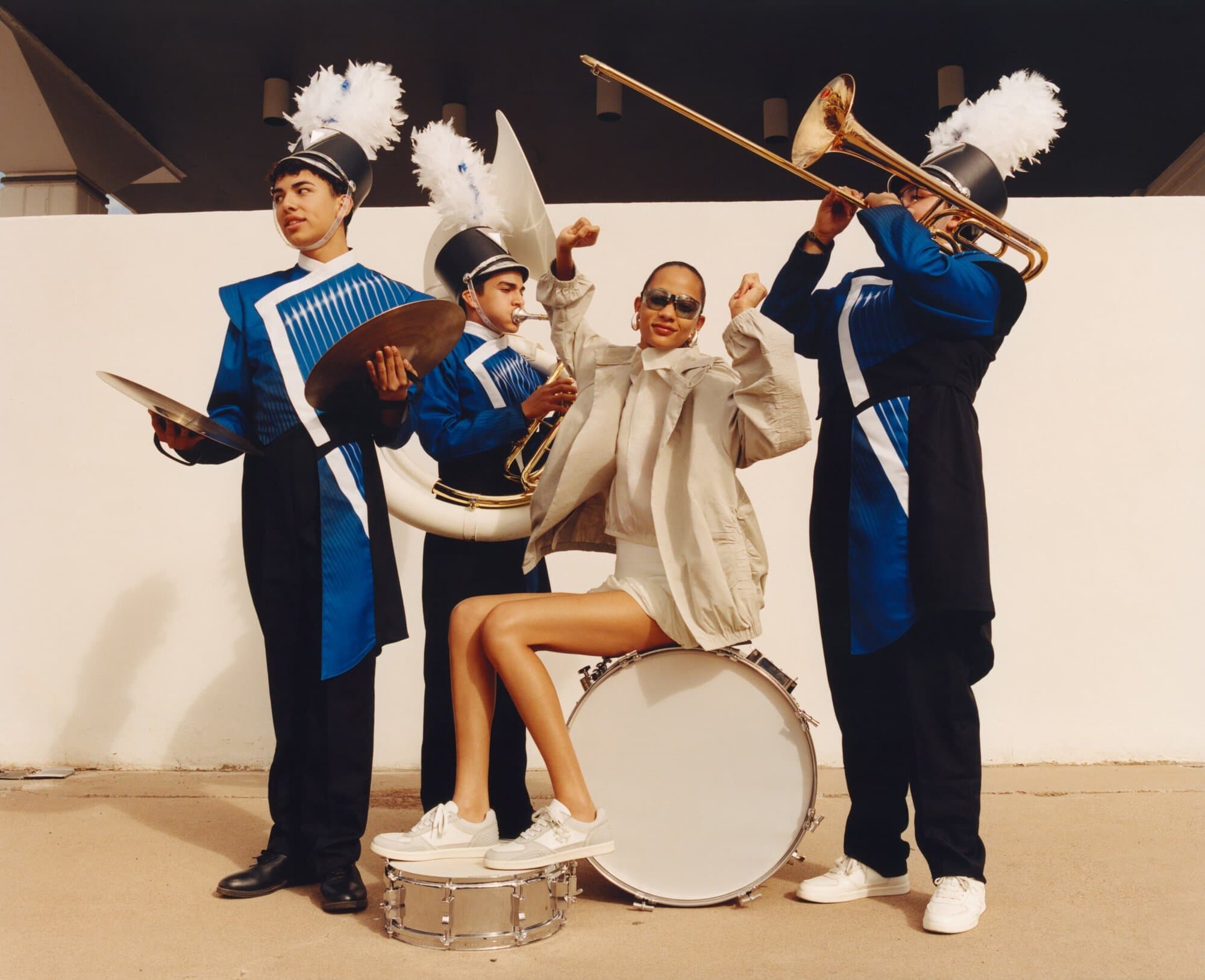 Group portrait of three marching band members in uniform with a model in sportswear, seated on a drum, smiling with her arms raised.