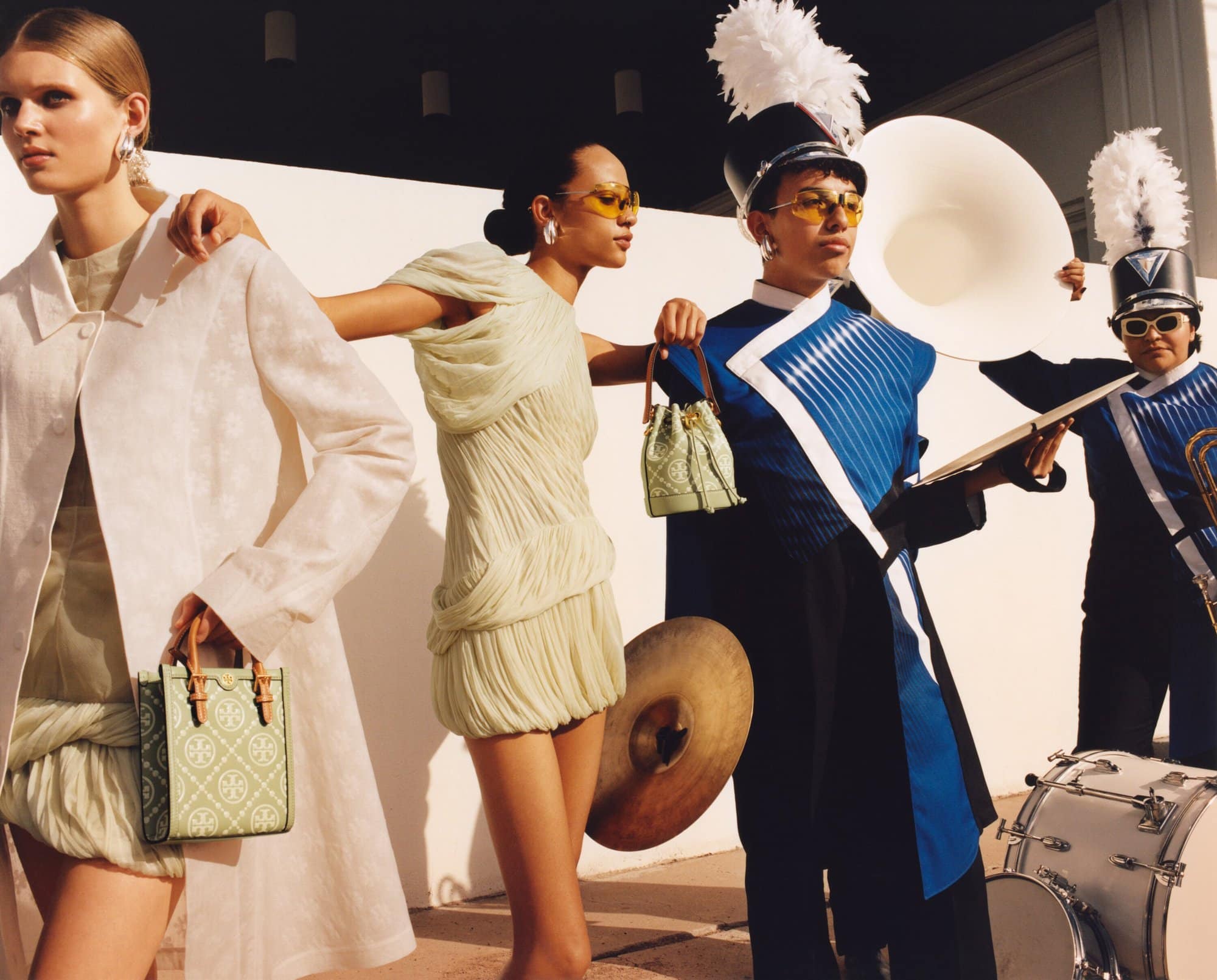 Fashion editorial featuring models in dresses and band members in uniform with brass instruments against a white wall backdrop.