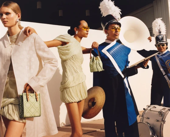 Fashion editorial featuring models in dresses and band members in uniform with brass instruments against a white wall backdrop.