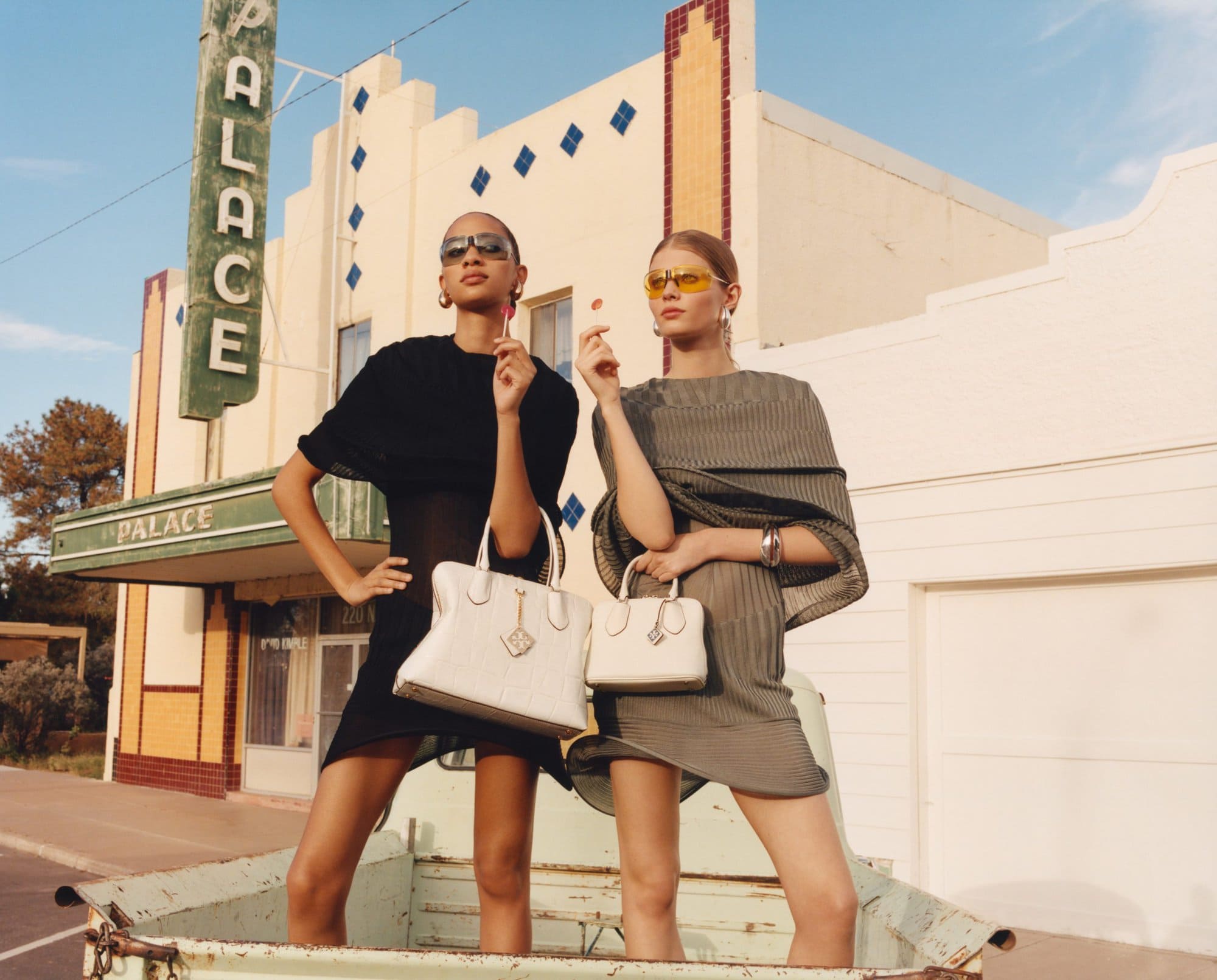 Two women pose outdoors by vintage Palace theater in coordinated retro dresses and handbags, holding lollipops, under a clear blue sky.