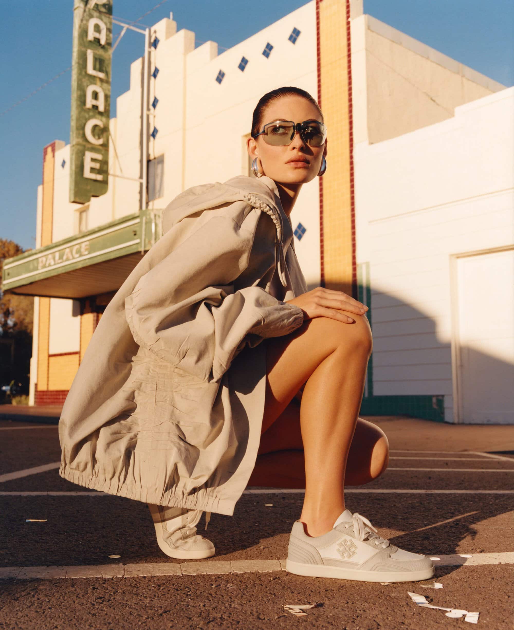 Model in sunglasses and a light jacket crouching outdoors near a pale building with a green Palace sign.