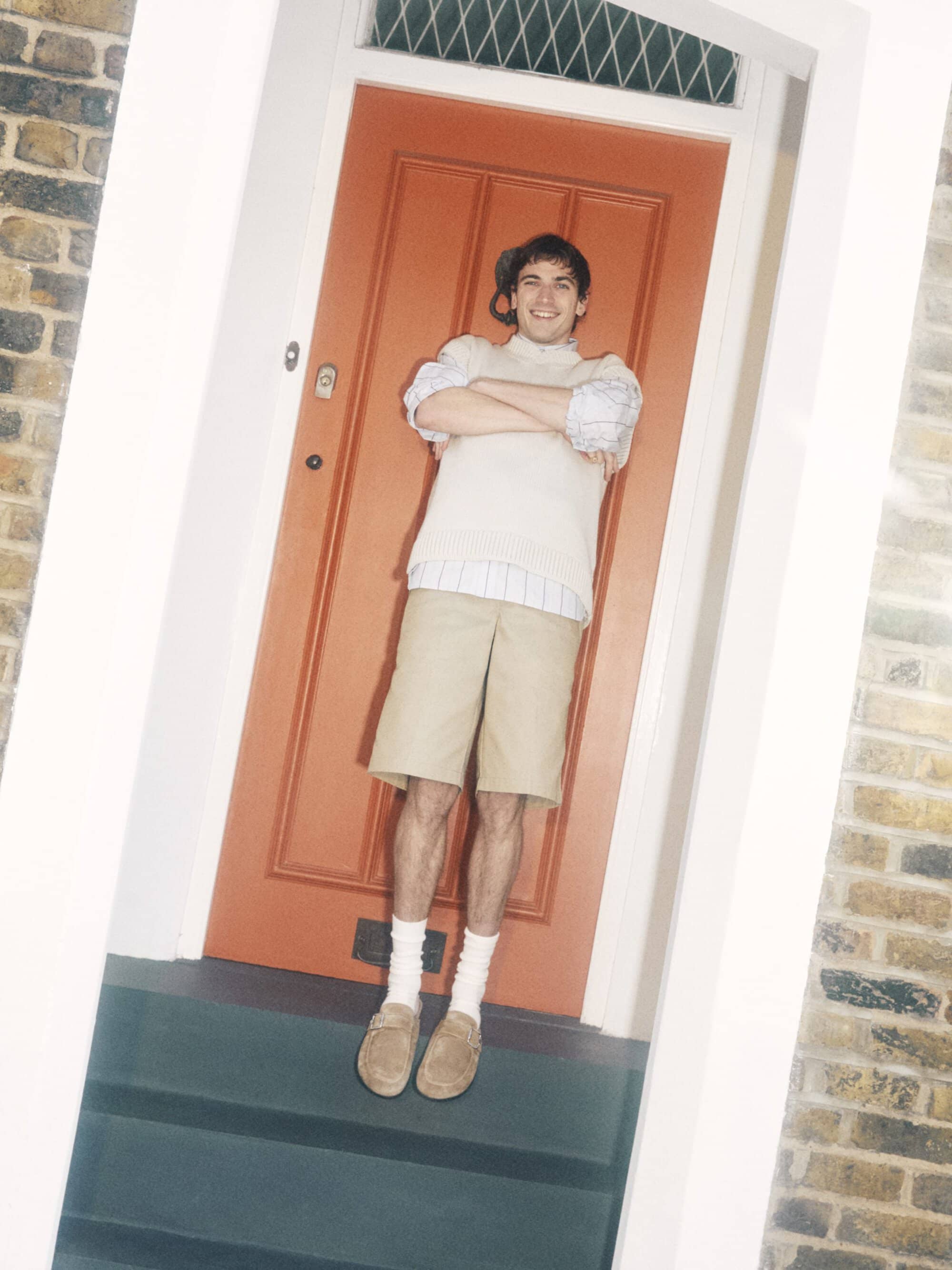 Young man in beige shorts and sweater standing in a doorway with orange door and brick wall in soft, diffused light.