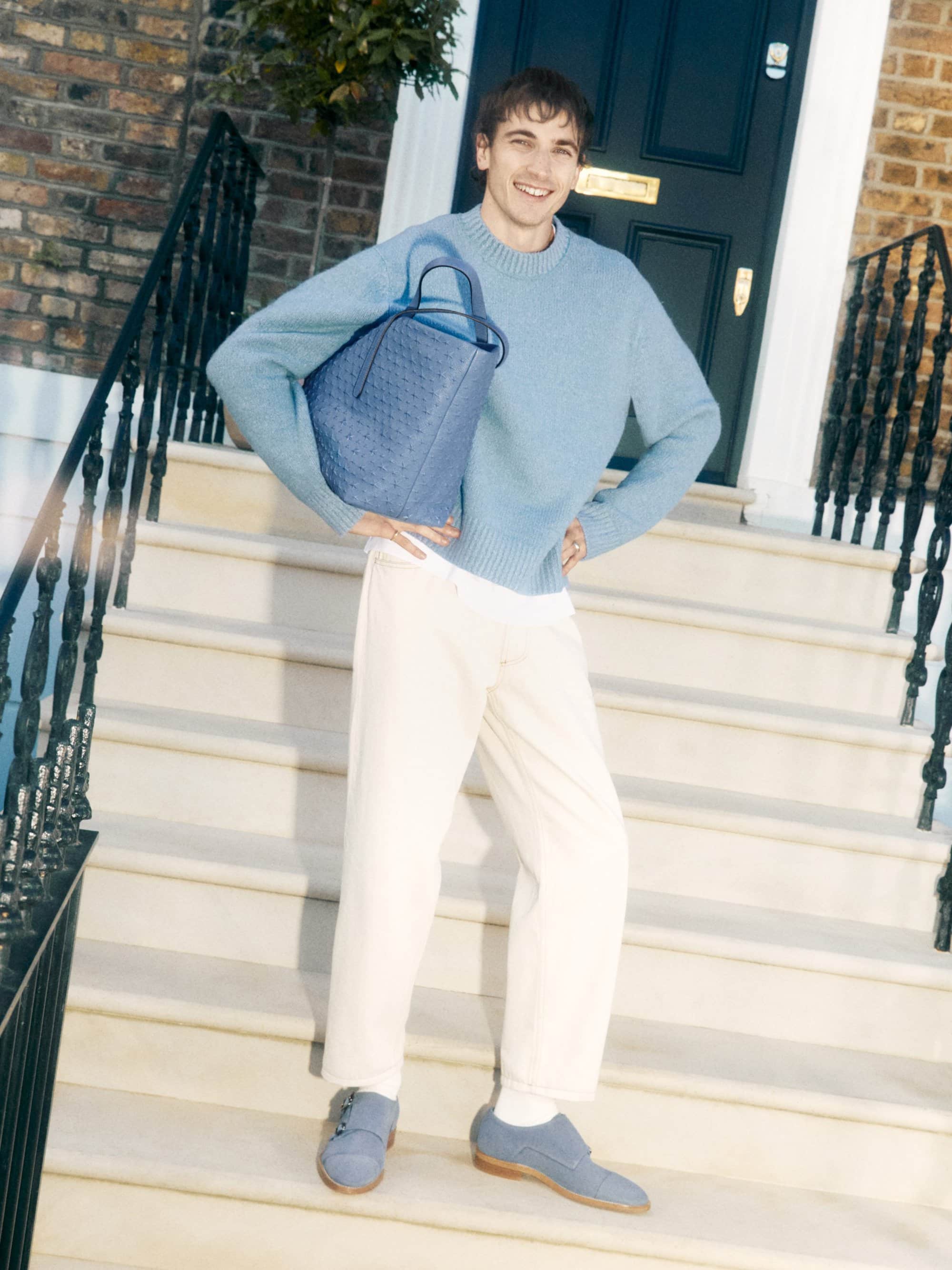 A young adult man in a blue sweater and cream pants poses on steps with a blue patterned tote bag outside a brick building.