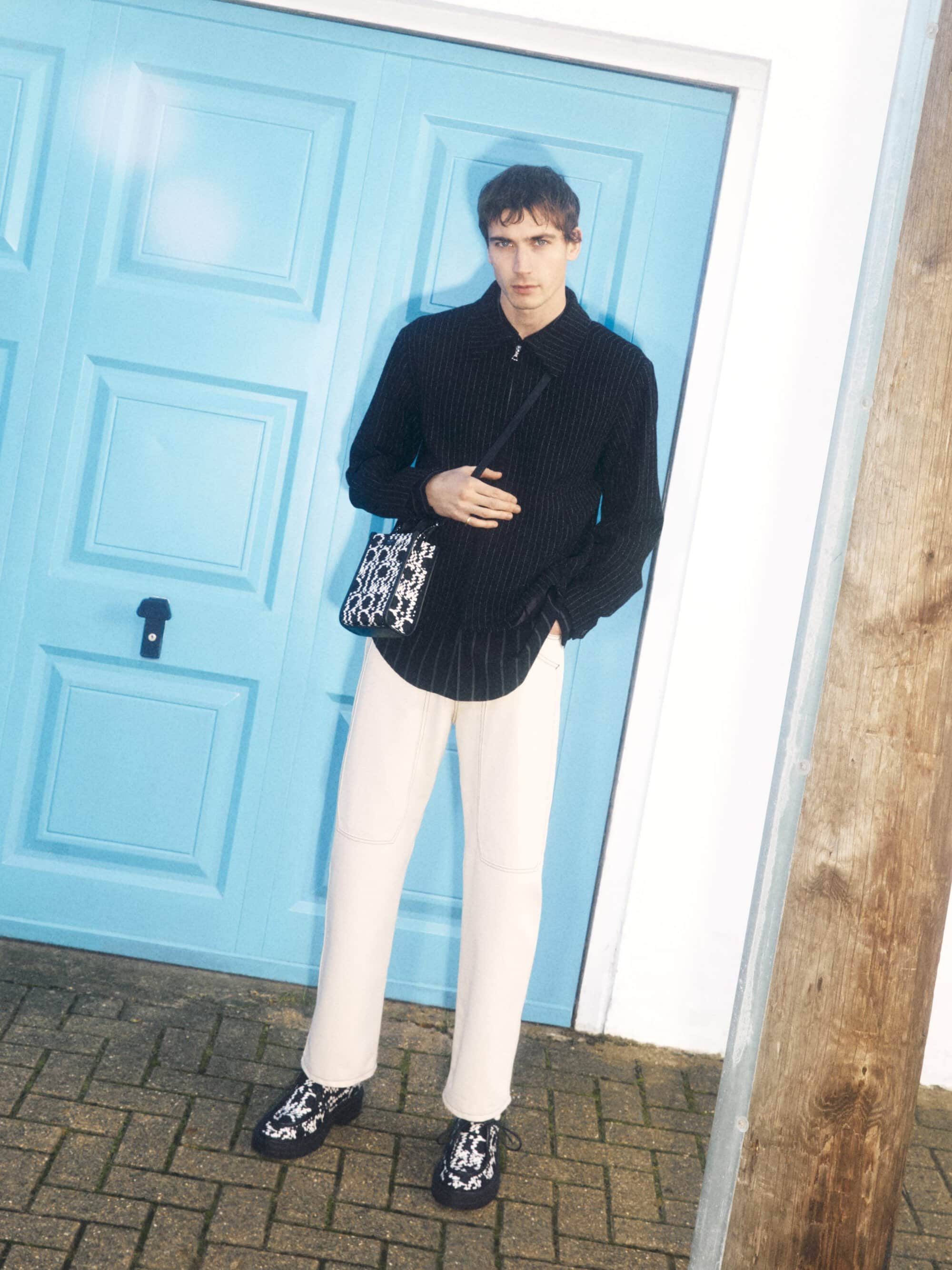 A full shot of a young adult man wearing a black pinstripe shirt, cream pants, and patterned shoes, posing against a light blue door outdoors.