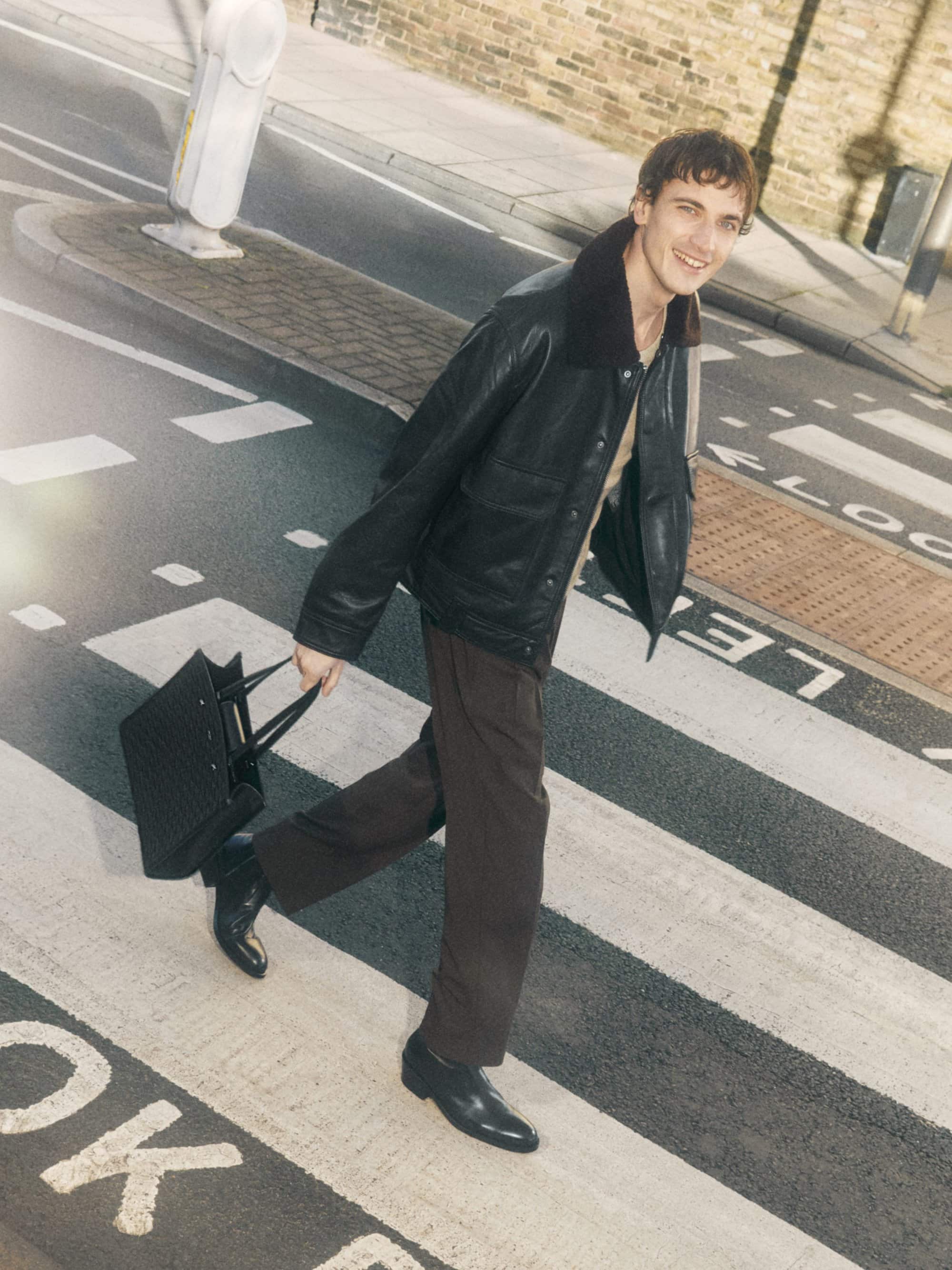 A young adult man crosses a pedestrian crosswalk in a leather jacket, pants, and dress shoes, carrying a black tote bag, smiling.