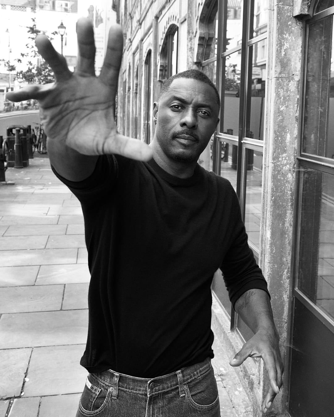 Black and white portrait of Idris Elba raising his hand toward the camera on an urban street.