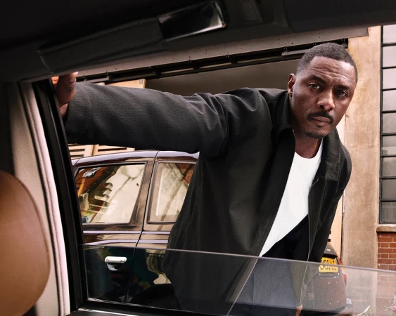 Idris Elba looking through car window. Black car with yellow license plate in background. Brick wall on the side of building.