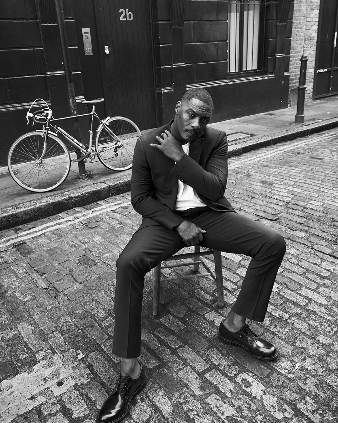 Black and white portrait of a seated, stylish adult male in a suit, posing outdoors with a bicycle and a building facade backdrop.