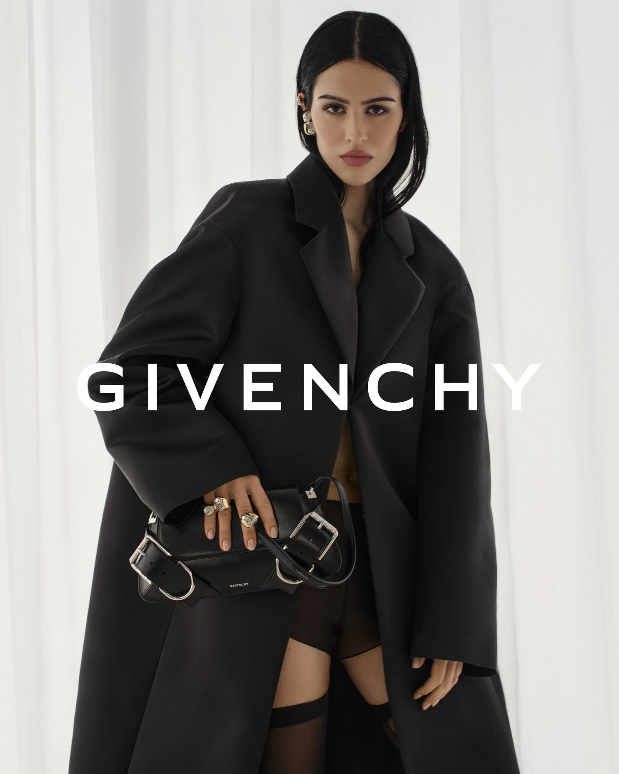 Woman wearing a black overcoat, jewelry, and holding a black handbag in front of a white backdrop, logo of Givenchy displayed.