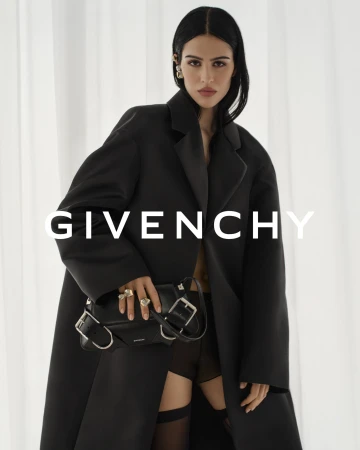Woman wearing a black overcoat, jewelry, and holding a black handbag in front of a white backdrop, logo of Givenchy displayed.