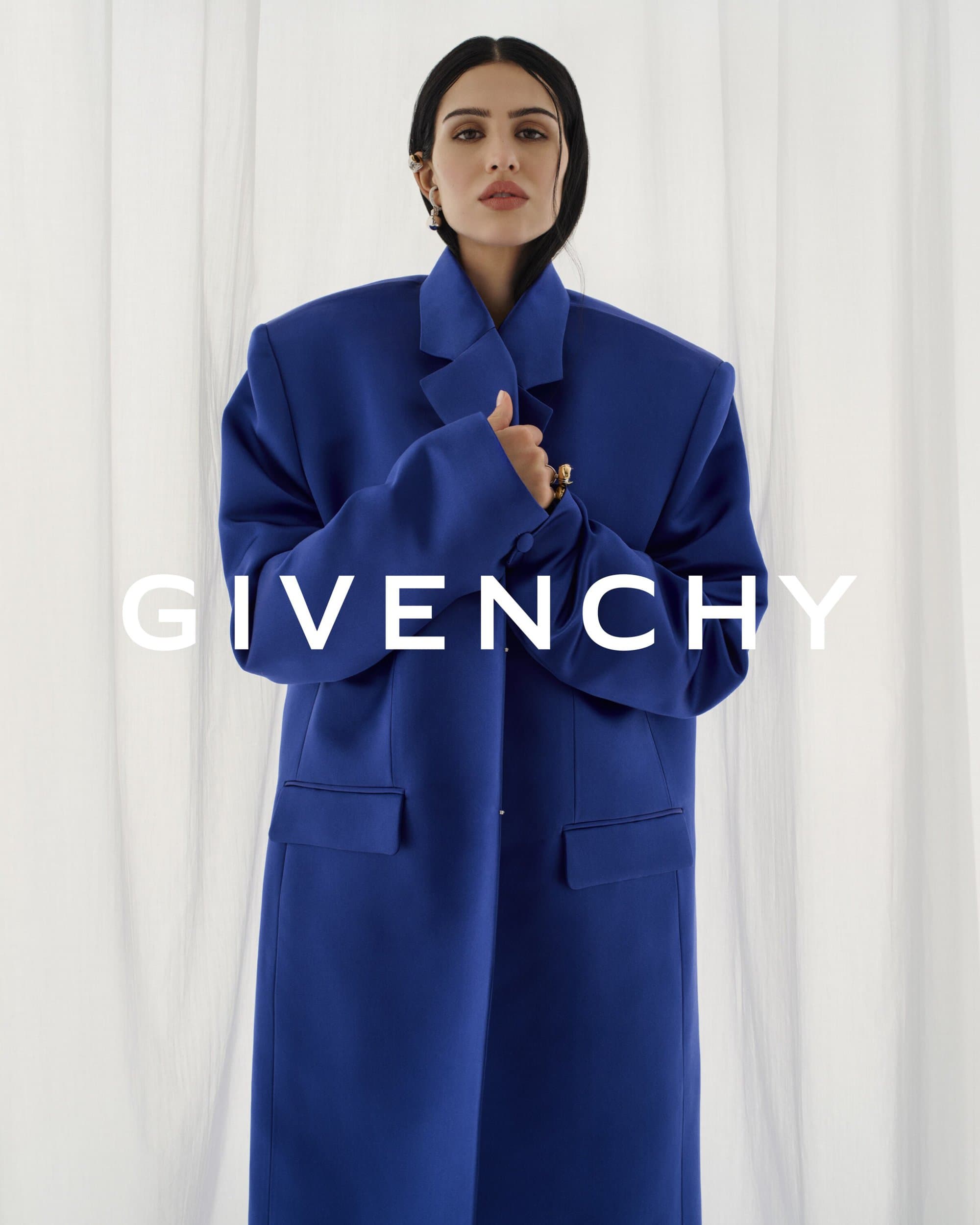 Fashion shot of a female model wearing a blue oversized coat standing against a white draped backdrop, featuring the GIVENCHY logo.