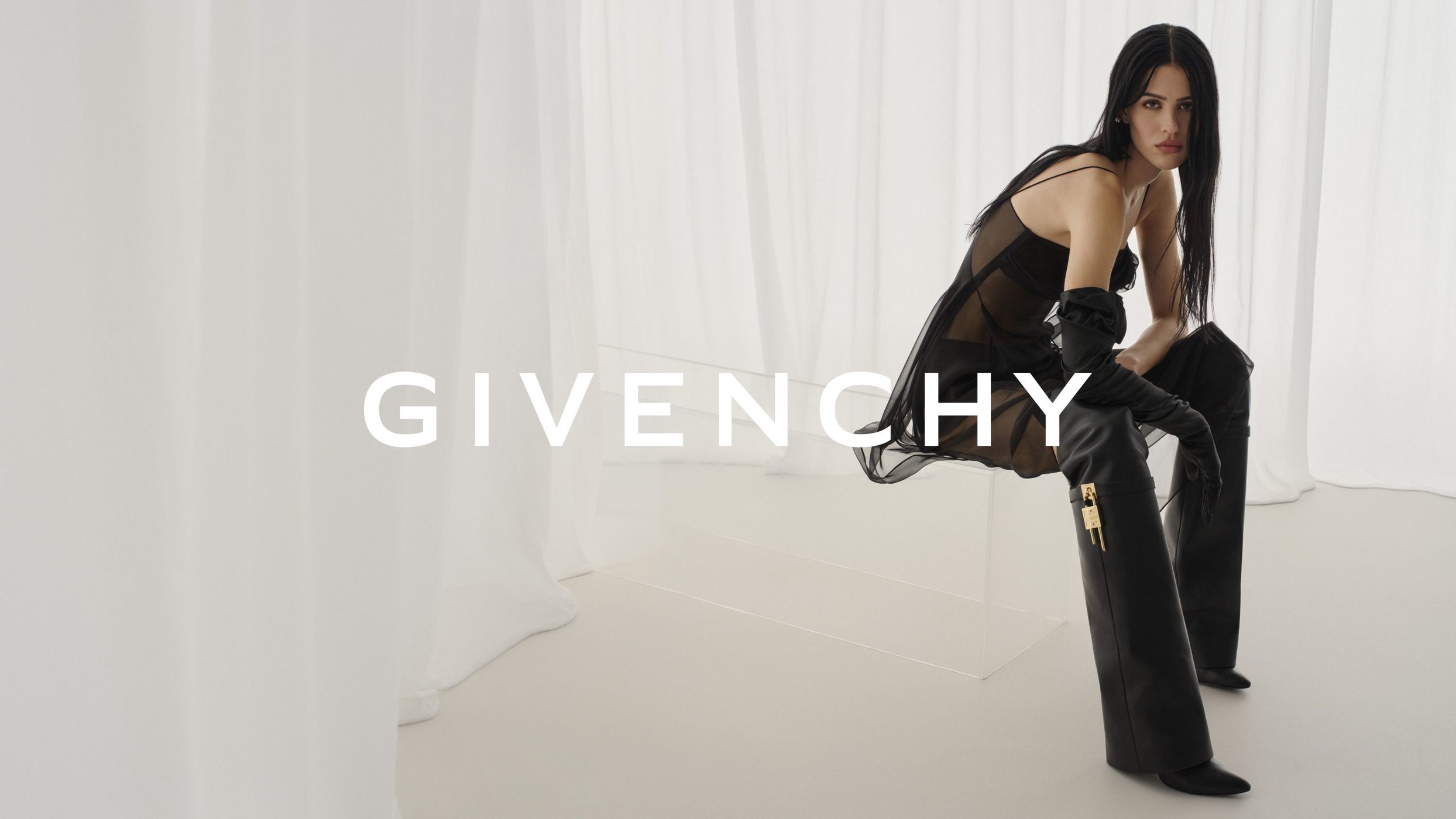 Fashion shoot of a woman sitting on a glass bench in a sheer black dress and thigh-high leather boots, Givenchy logo in the foreground.
