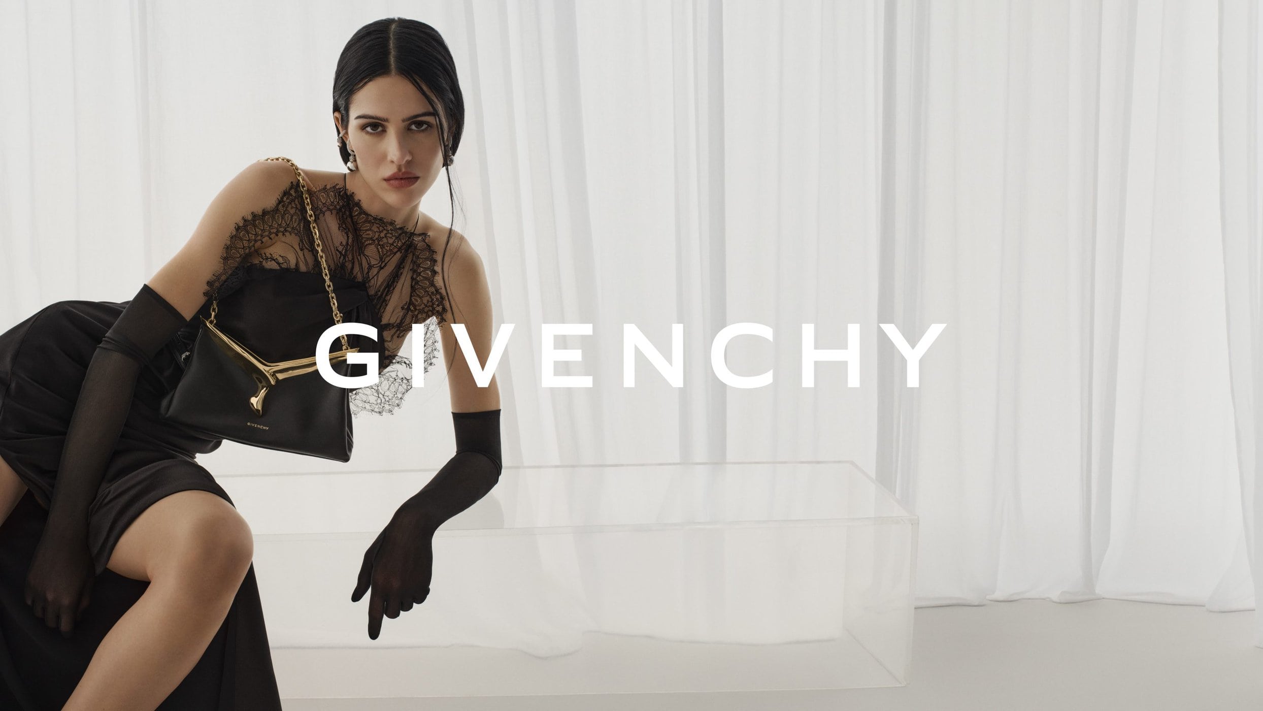 Fashion shot of a woman in black dress with lace detail holding a purse, posing in front of white curtains and a glass table.