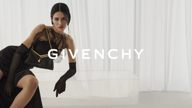 Fashion shot of a woman in black dress with lace detail holding a purse, posing in front of white curtains and a glass table.