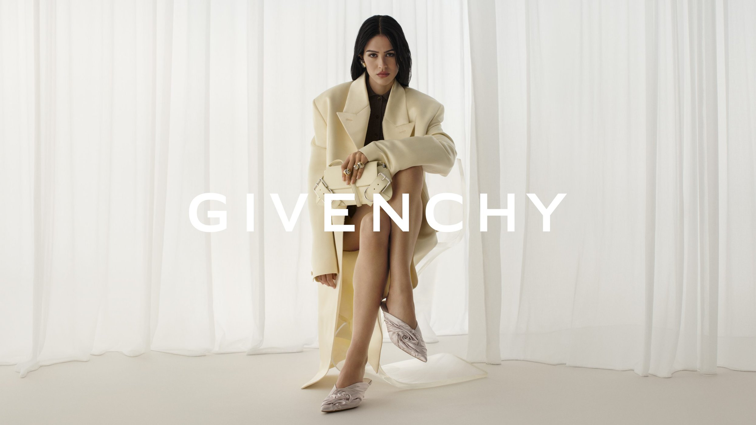 Editorial fashion shot of a woman in an oversized coat holding a purse, seated against a draped curtain backdrop, featuring a logo.
