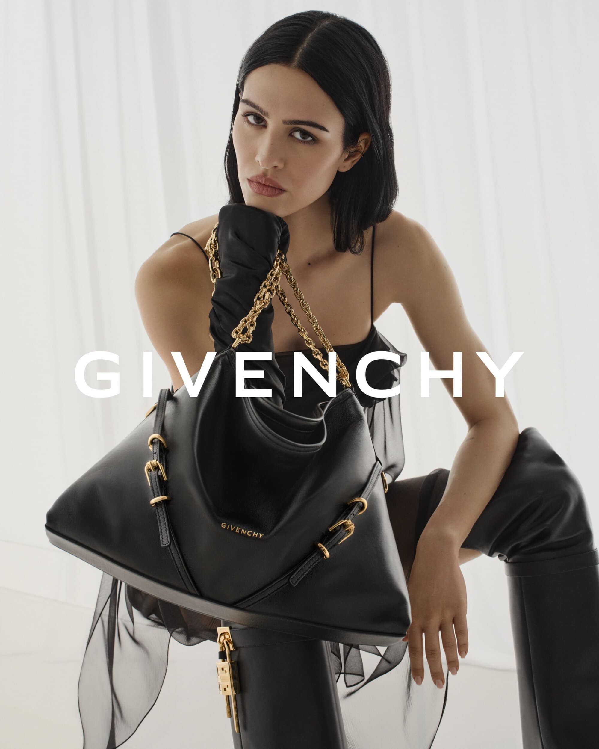 Elegant brunette model poses with a black Givenchy handbag, wearing a black dress and gloves in a bright, high-key fashion shot.