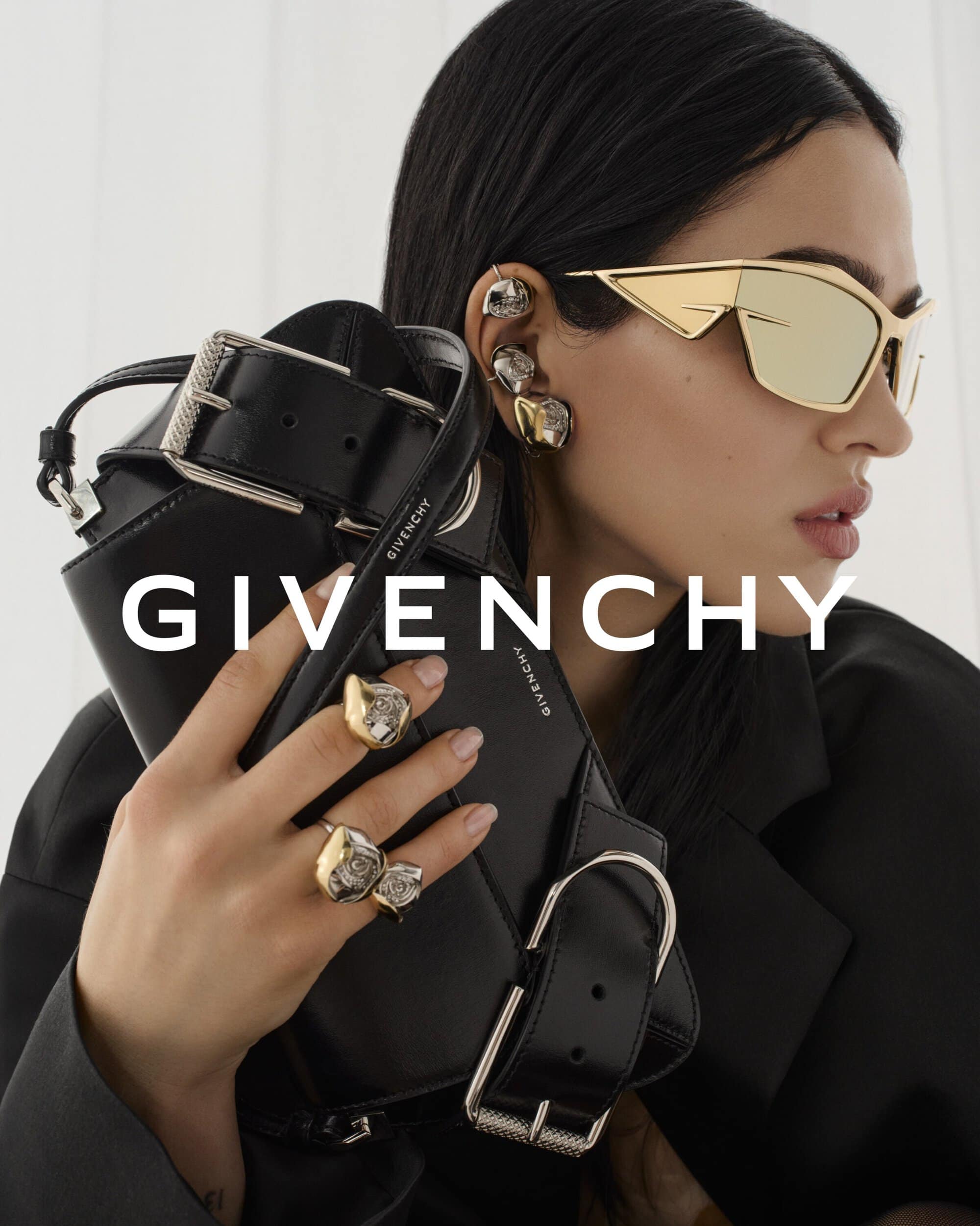 Fashion advertisement featuring a model with statement sunglasses and rings, holding a black handbag, against a bright white background.