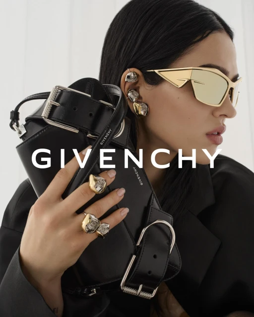 Fashion advertisement featuring a model with statement sunglasses and rings, holding a black handbag, against a bright white background.