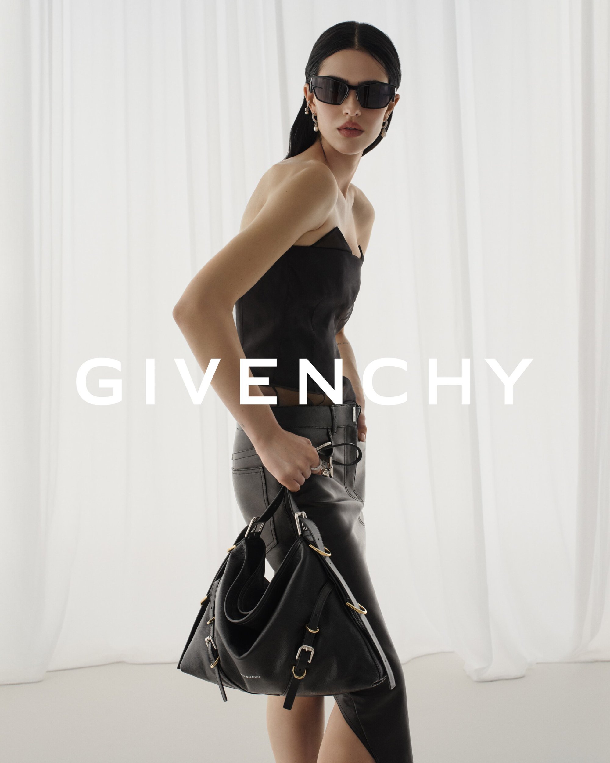 Brunette woman in a black top and leather skirt poses with a Givenchy handbag against a light background in a fashion editorial shot.