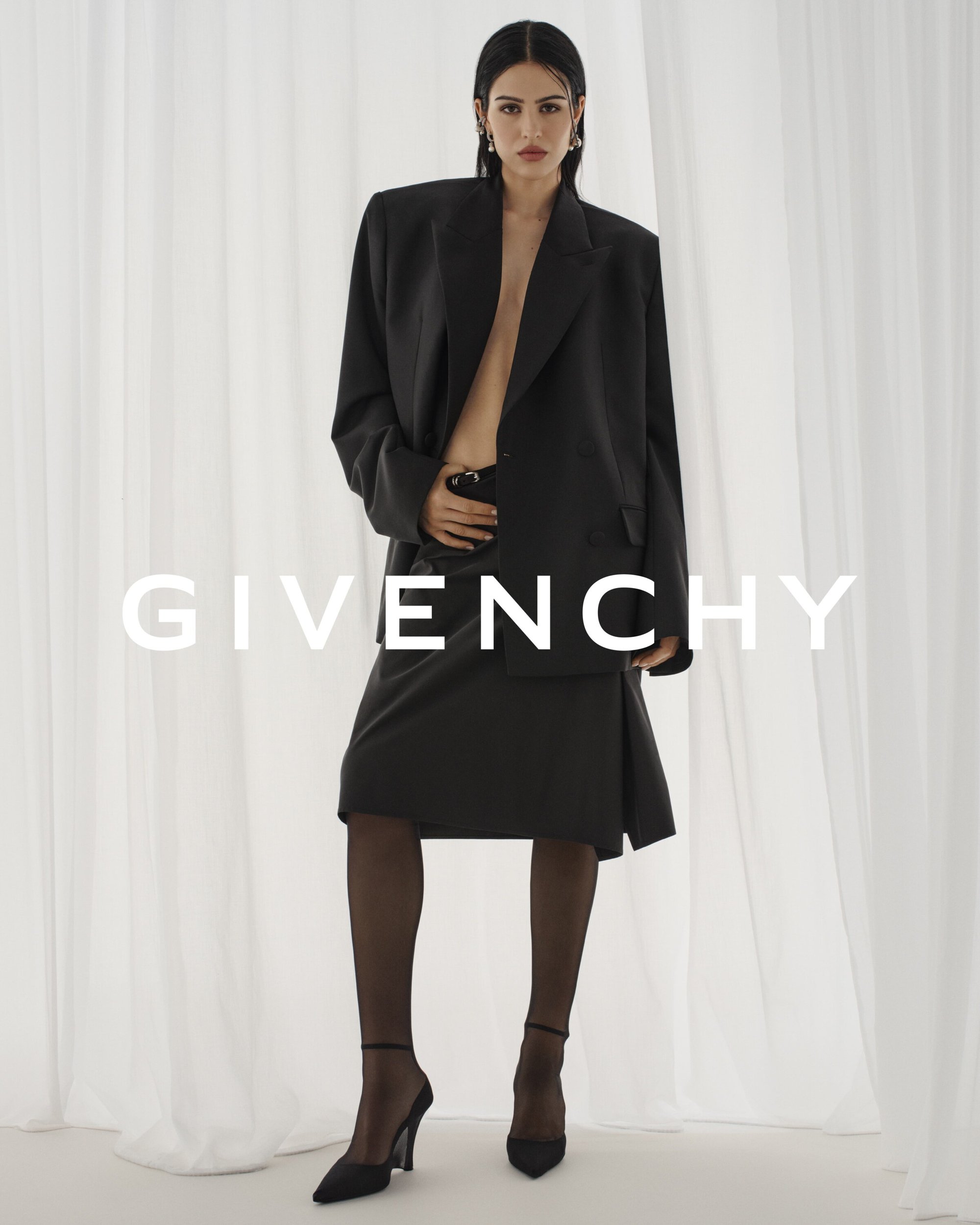 Woman in oversized black blazer and skirt standing in front of white curtains with the Givenchy logo in center frame.