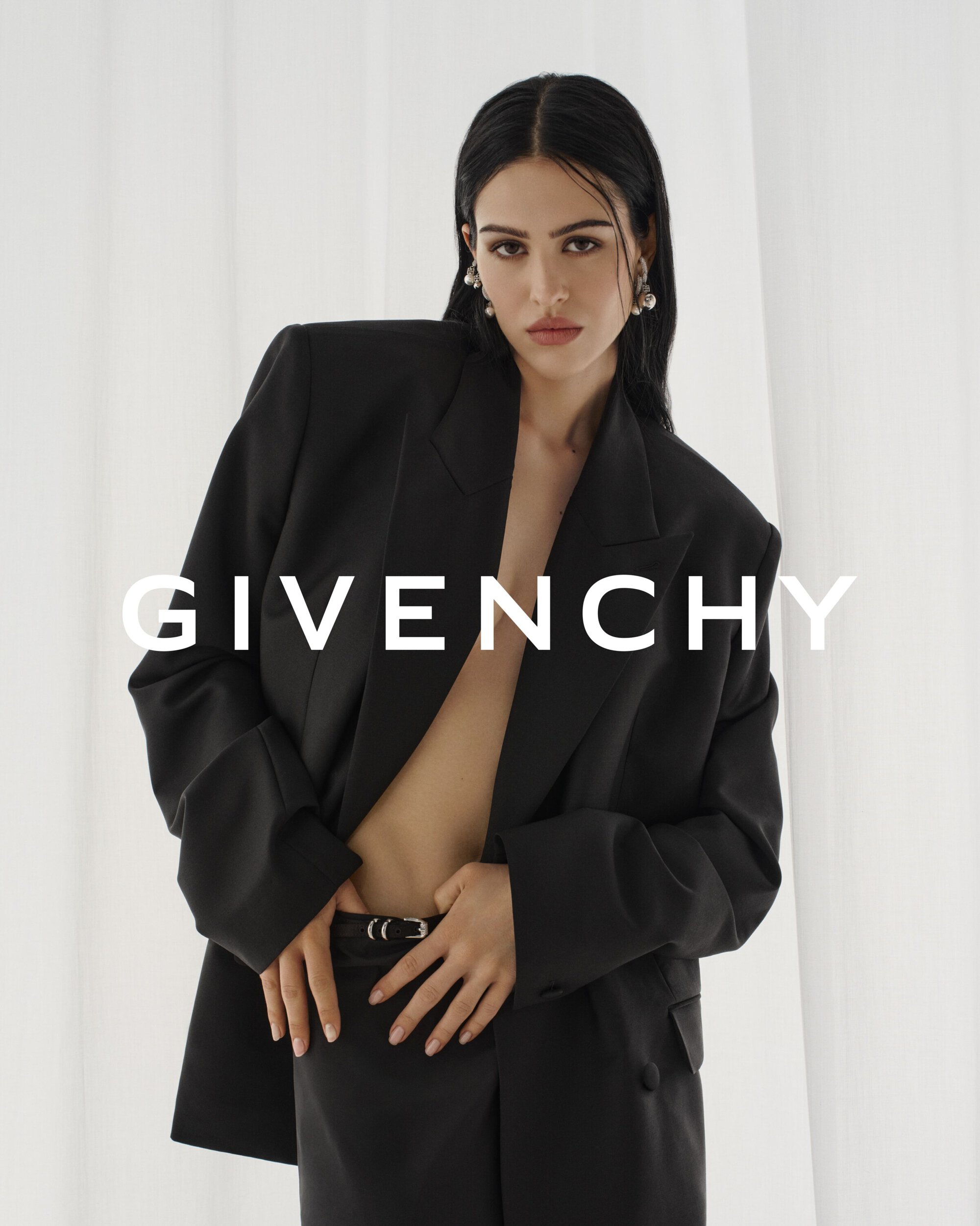 A woman in a black blazer poses in front of a white background, hands resting on a black belt, Givenchy logo visible.