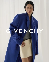 Model wearing a royal blue oversized blazer over a grey top and cream skirt against a white curtain background.