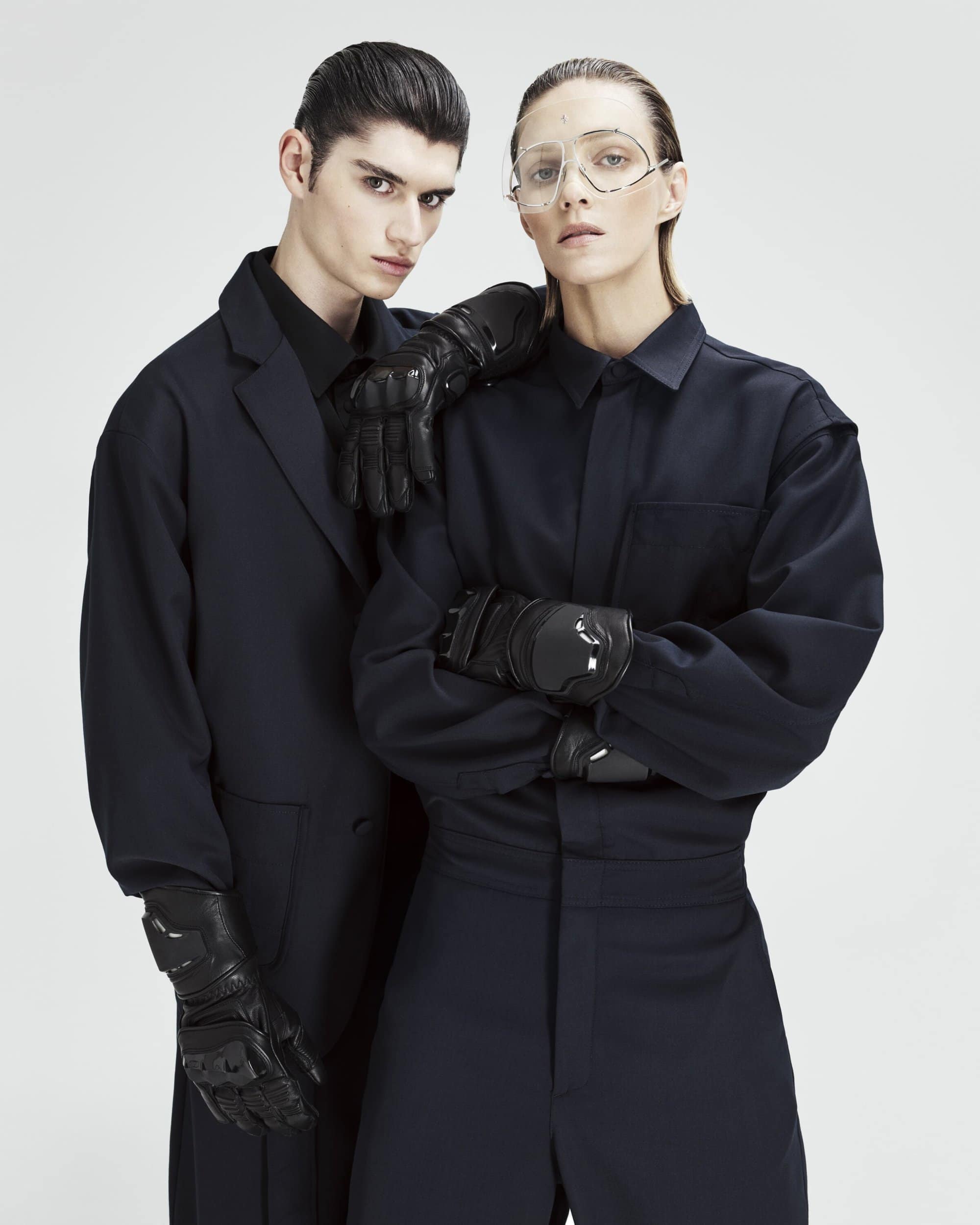 Two fashion models in dark blue outfits and black leather gloves pose in a studio, showcasing modern style and edgy sophistication.