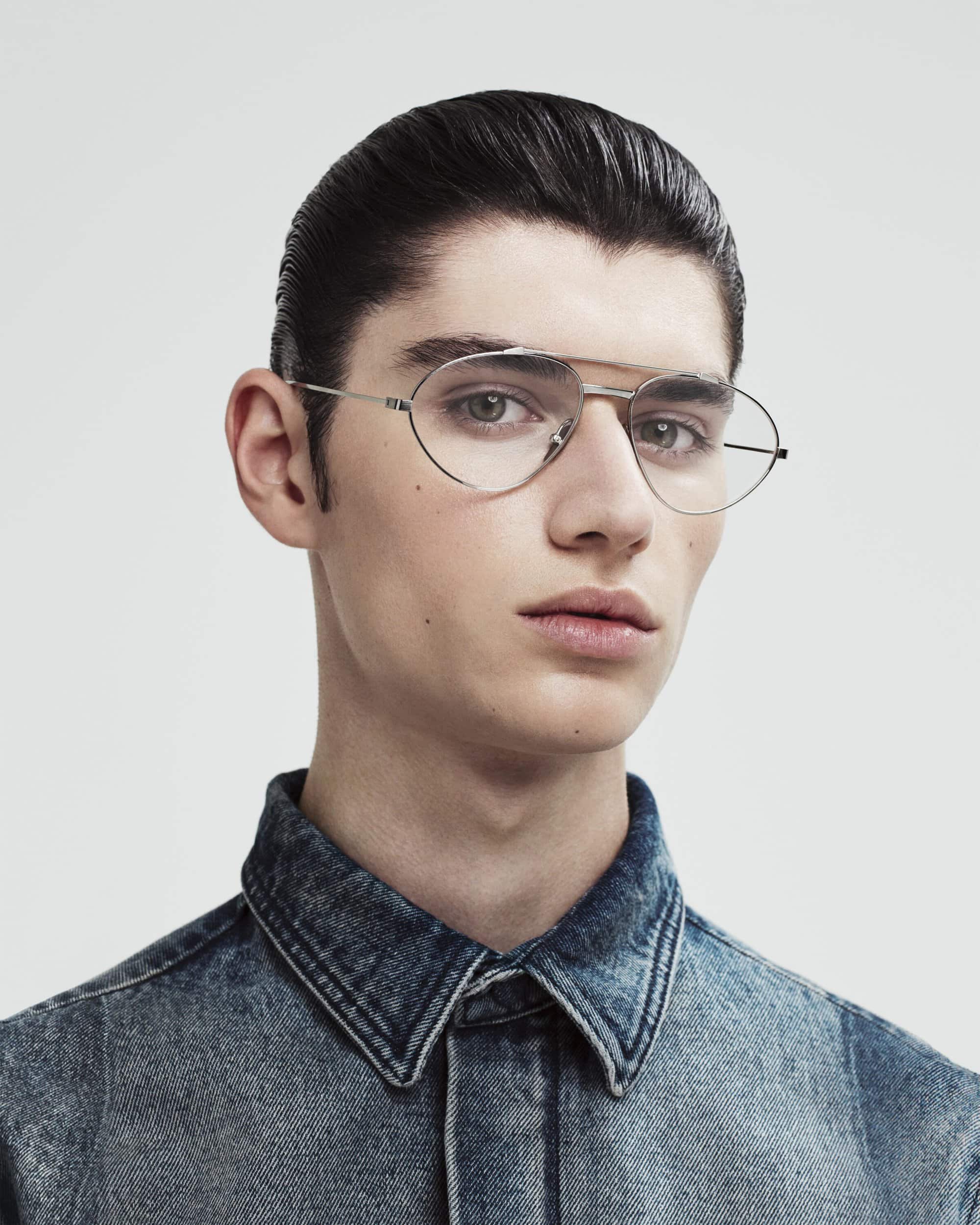 Close-up of a young adult male with dark hair styled back, wearing wire-frame glasses and a denim shirt, against a neutral background.