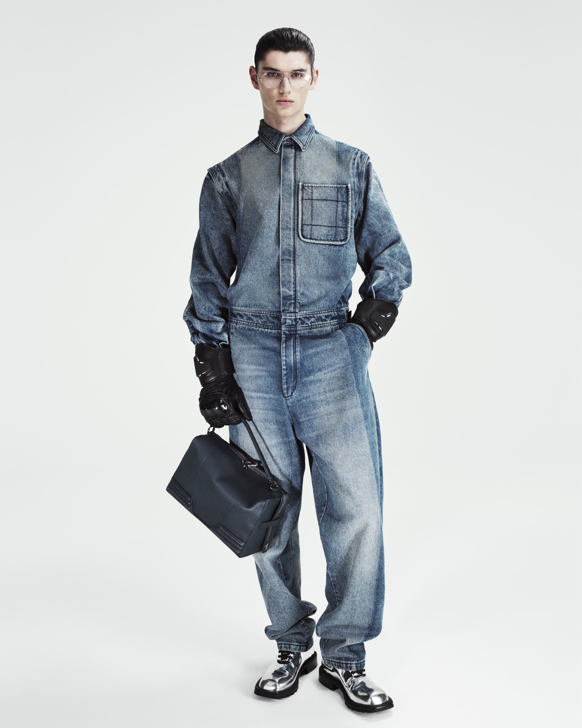 Full-length shot of a male model in a denim jumpsuit holding a black bag, accessorized with gloves and metallic shoes in a white studio.