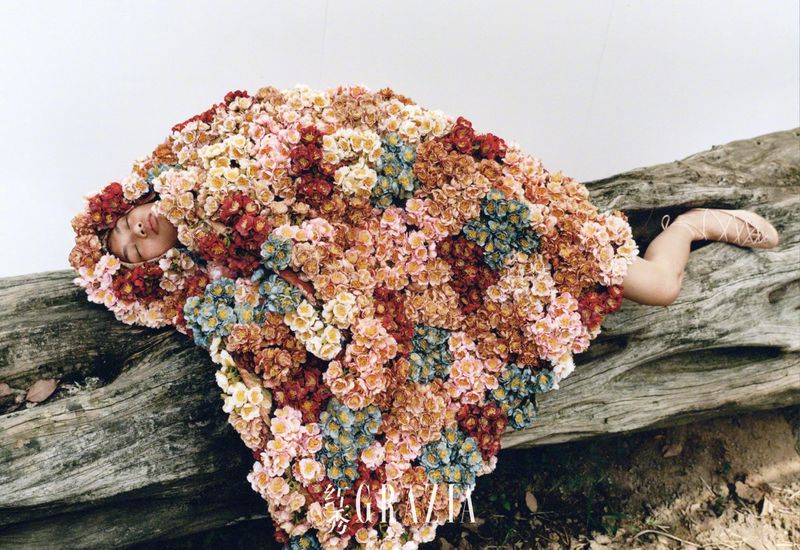 Woman covered in colorful flowers, lying on a weathered log in an artistic floral arrangement, against a plain white background in a studio.