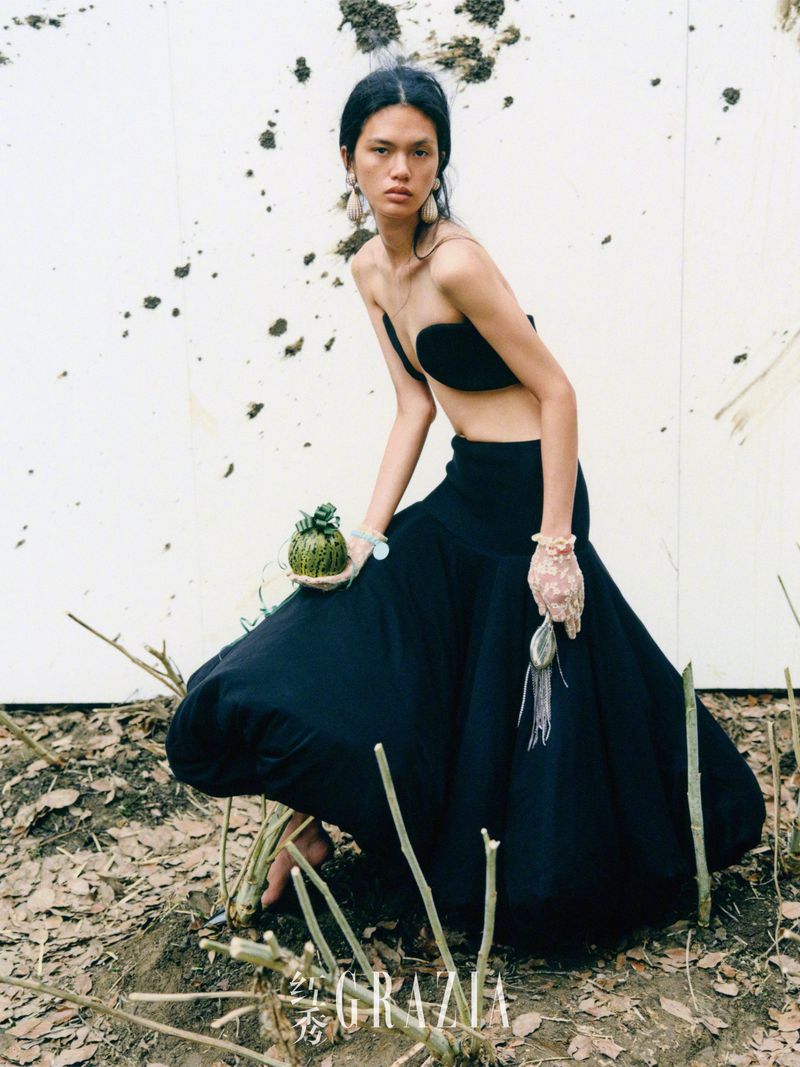 A woman in a black two-piece outfit and long skirt poses in a dirt field in front of a white wall, holding a green, moss-covered ball.