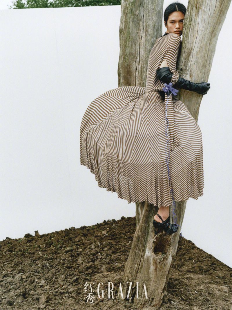 Striking editorial fashion shot of a model in a striped dress perched on a tree, set against a white backdrop and earthy foreground.