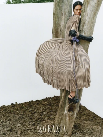 Striking editorial fashion shot of a model in a striped dress perched on a tree, set against a white backdrop and earthy foreground.