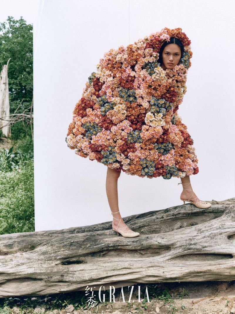 Fashion editorial shot featuring an Asian woman in a large floral garment, standing on a log against a white backdrop with a natural setting.