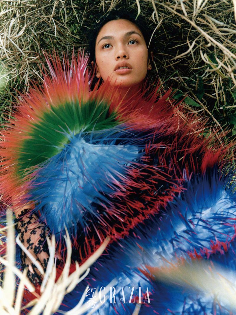 Fashion photo of a woman lying in grass wearing a feathery blue, green, and red dress with the title GRAZIA visible in the foreground.