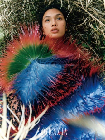 Fashion photo of a woman lying in grass wearing a feathery blue, green, and red dress with the title GRAZIA visible in the foreground.