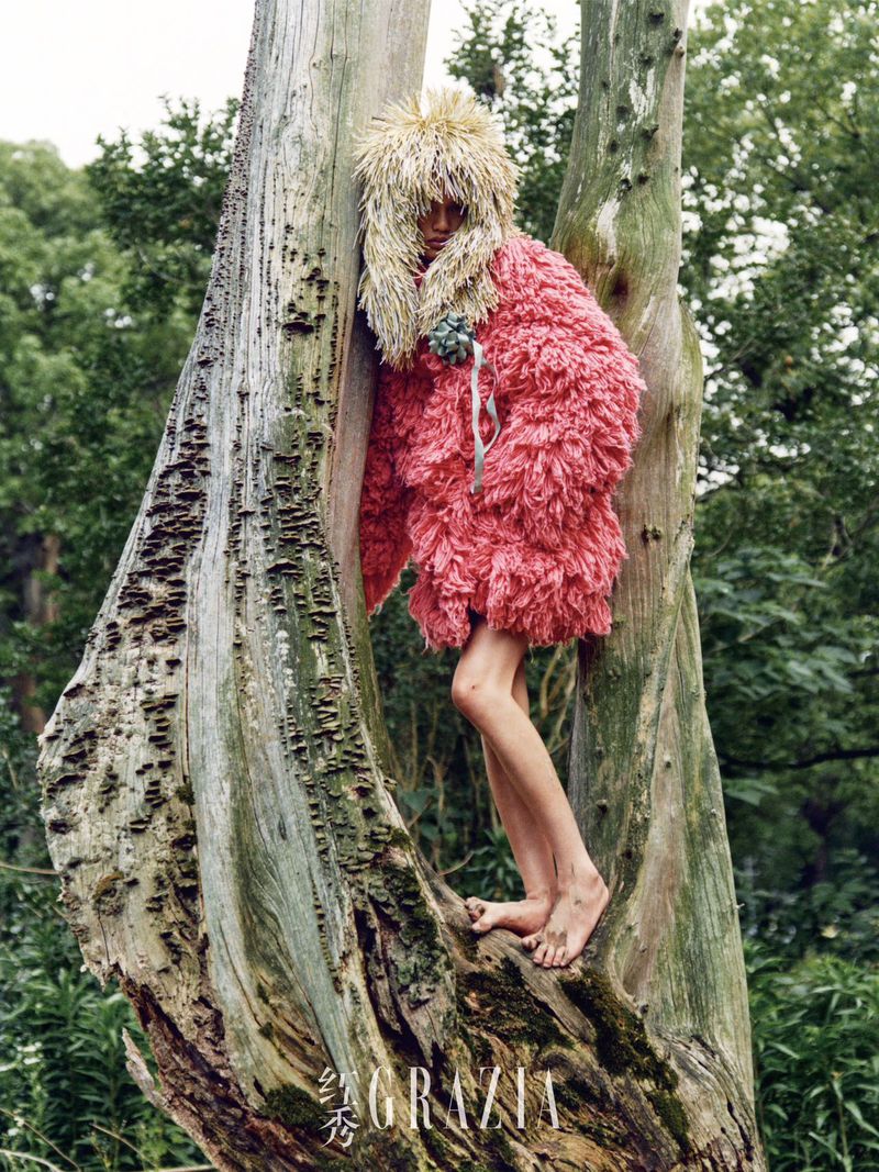A bare-footed androgynous model poses wrapped in the hollow of a mossy tree, wearing a furry pink coat and shaggy blond wig in nature.