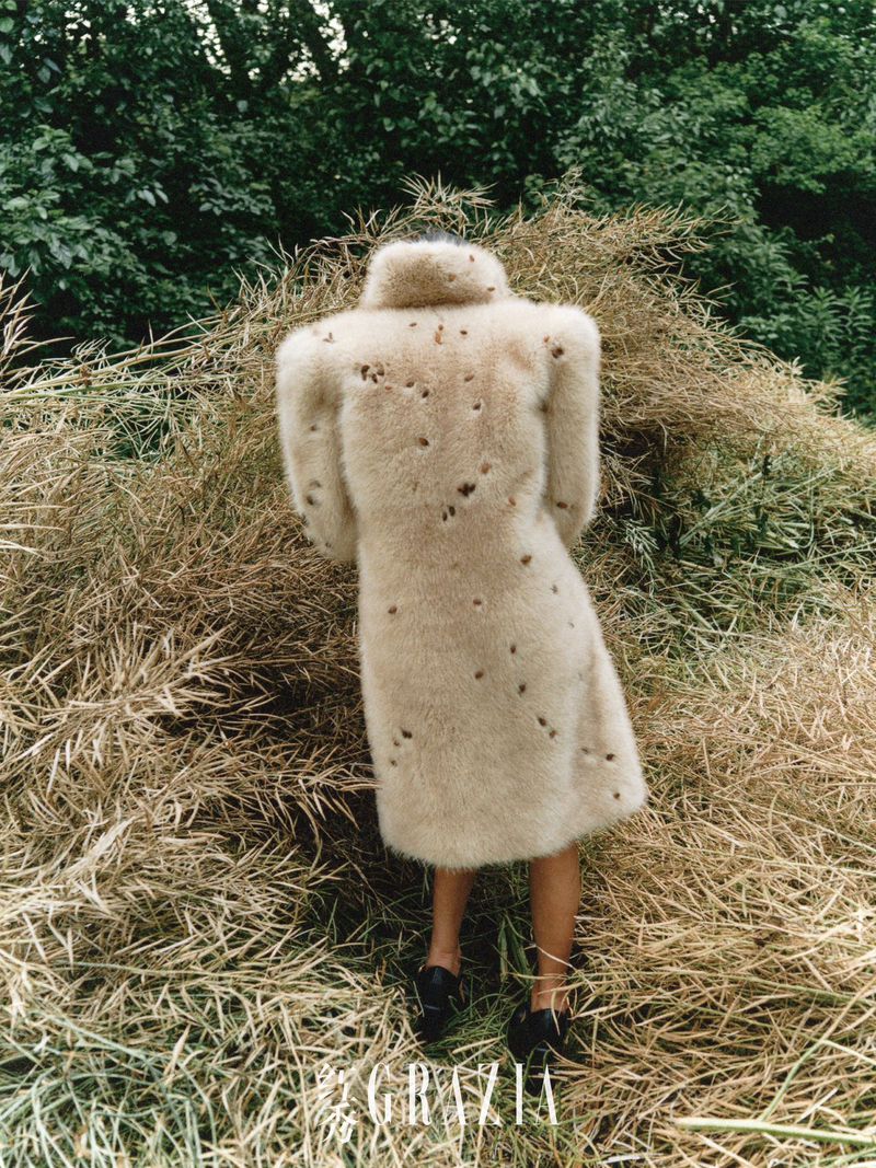 Woman in a textured coat stands in a field of golden hay, back to camera, fashion editorial shot, outdoor setting, natural light, style concept.