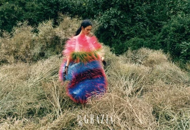 Model Qun Ye wearing a colourful feathered avant-garde dress amidst wild tall grass and overgrown vegetation.