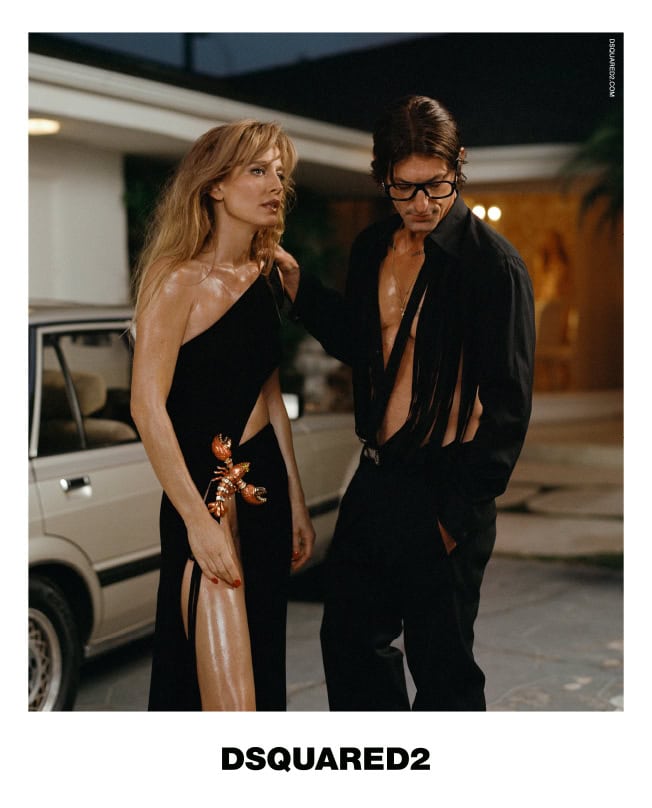 A woman in a black dress poses with a man in a black suit in a fashion editorial, bathed in soft lighting near a vintage car.