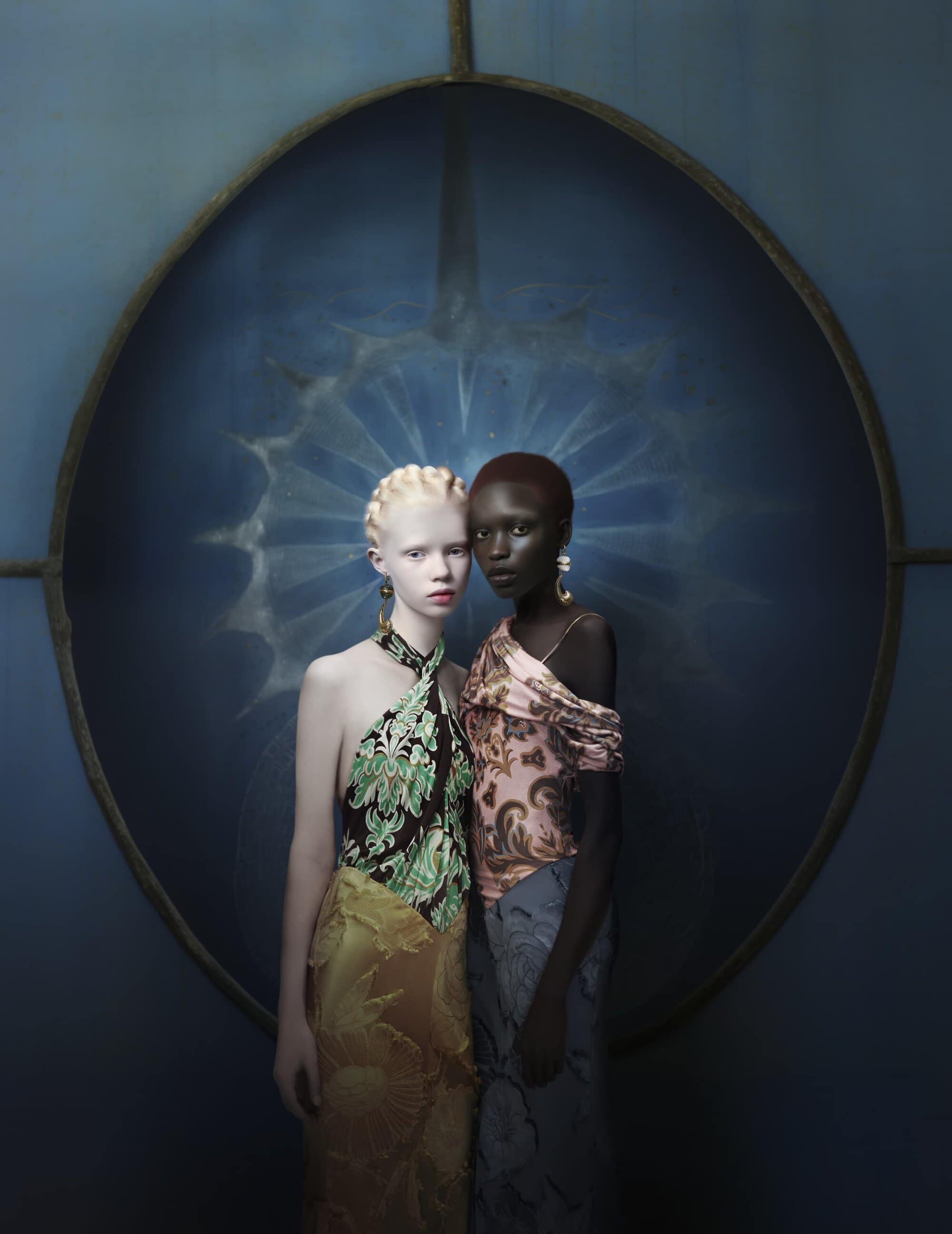 Two women pose in a fashion shot against a blue backdrop. The woman on the left has albinism, and the woman on the right is Black.