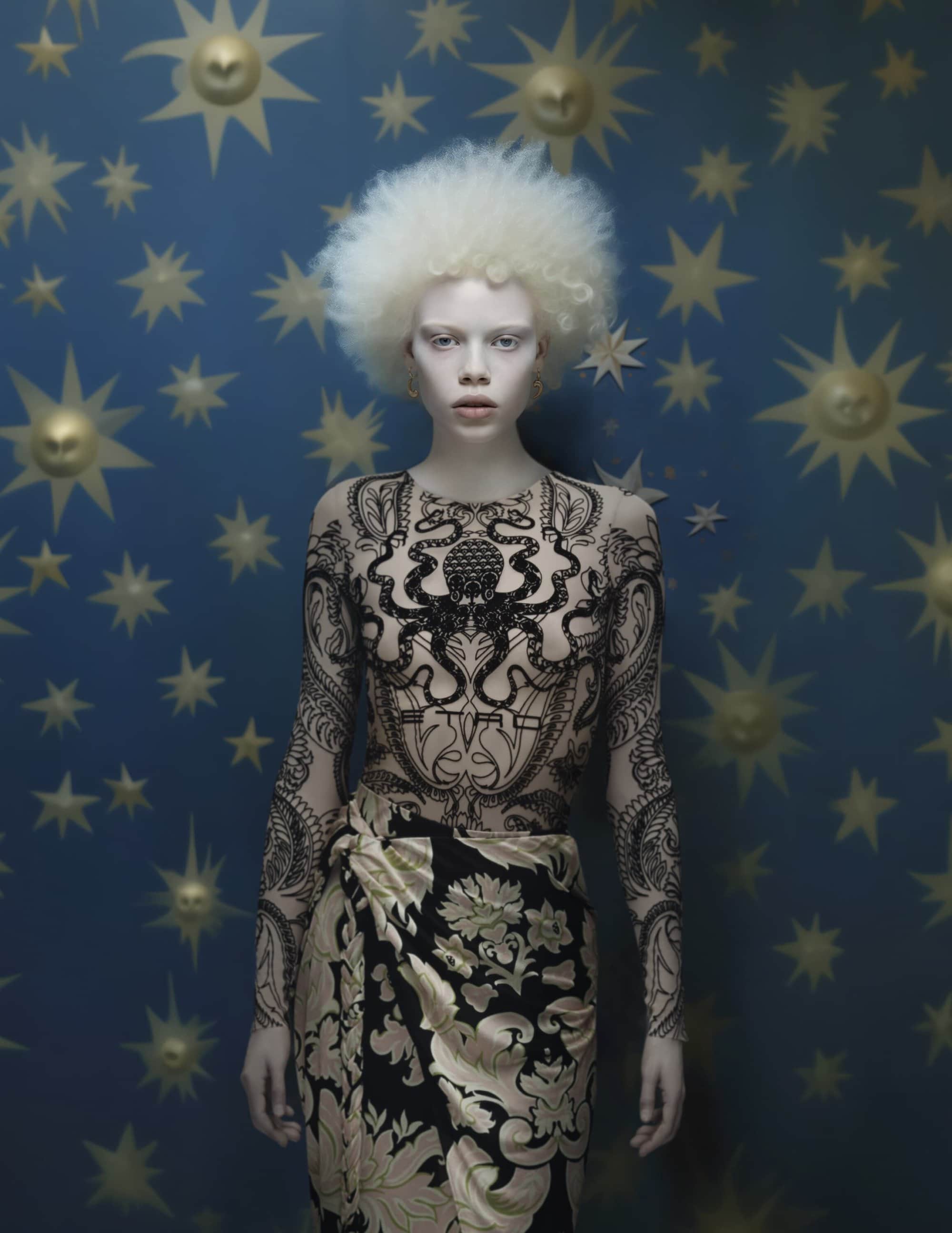 Albino female model in patterned bodysuit and skirt posing against a blue celestial backdrop with golden sun and star shapes. Fashion portrait.