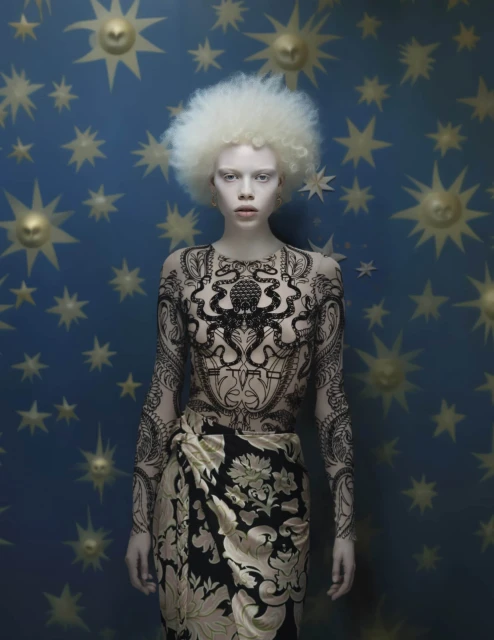 Albino female model in patterned bodysuit and skirt posing against a blue celestial backdrop with golden sun and star shapes. Fashion portrait.